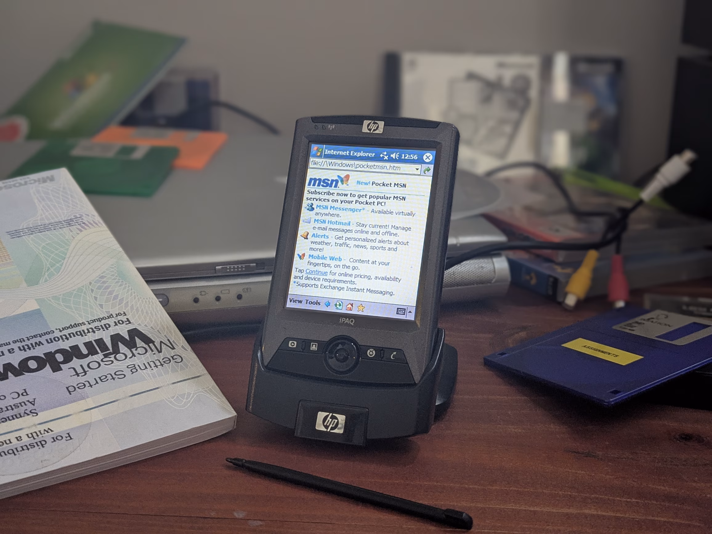 A grey PDA in a dock open to a Pocket MSN subscription page in Internet Explorer. The browser has an address bar at the top and a menu at the bottom. The operating system menu can be seen at the very top, and the entire interface is Luna themed. In the background is 2000s tech miscellany.